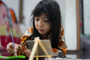 A young girl focuses while painting on a small canvas using a brush. She wears a colorful batik outfit as she works creatively at home, showcasing a genuine moment of childhood art and imagination.