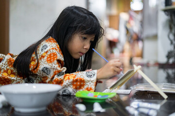 A young girl wearing a patterned traditional shirt is painting carefully on a canvas indoors. She is using a small brush and focusing on her creative artwork. 