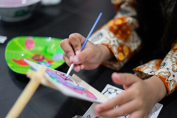 A close-up of a child’s hand painting colorful artwork with a small brush on a canvas. The scene shows bright colors and focused artistic activity.
