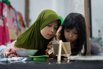 Two young girls painting together on a small easel while lying on the floor at home. They look focused and happy as they share a creative activity using brushes and bright colors.