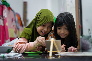 Two young girls painting together on a small easel while lying on the floor at home. They look focused and happy as they share a creative activity using brushes and bright colors.