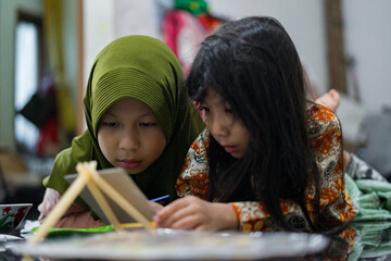 Two young girls smiling while painting on a small canvas. They lie on the floor and enjoy a fun creative moment using brushes and bright colors in a cozy indoor setting.