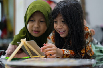 Two young girls smiling while painting on a small canvas. They lie on the floor and enjoy a fun creative moment using brushes and bright colors in a cozy indoor setting.