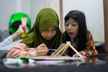 Two young girls smiling while painting on a small canvas. They lie on the floor and enjoy a fun creative moment using brushes and bright colors in a cozy indoor setting.