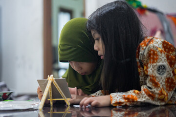 Two young girls are lying on the floor while painting together using a small easel and watercolor palette. One girl wears a green hijab, and the other has long hair and batik clothes.