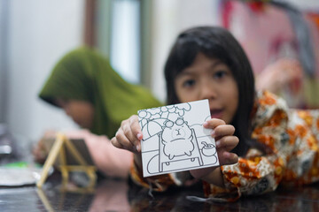 A young girl in batik clothes shows a small drawing board with a cute cartoon picture while lying on the floor. Another child in a green hijab is painting in the background.