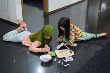 Two young girls are lying on the floor while painting and coloring. The girl in the green hijab focuses on her canvas, while the other girl smiles and watches closely. 