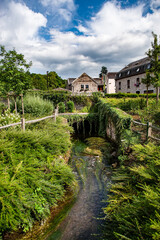 A charming stream winds beneath a lush green archway near the historic Spontin Castle in Belgium, surrounded by countryside vegetation and traditional stone buildings under a partly cloudy summer sky.