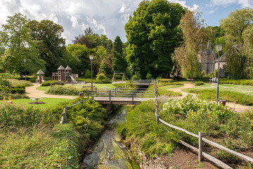 Lush garden landscape near Spontin Castle in Belgium featuring wooden bridges, flowering bushes, tall trees, and a playground under a partly cloudy sky creates a peaceful and charming countryside retr