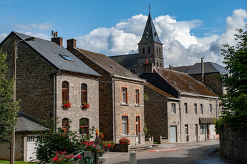 Stone houses with red flower boxes line a quiet street in the village near Château de Spontin in Belgium, with a historic church tower rising above under a bright blue sky with dramatic white clouds.