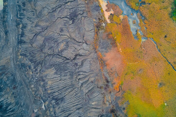 Abstract drone shot sharp boundary between two distinct landscapes: heavily eroded or industrial gray soil with intricate textures and brightly colored yellow, orange, and green marshland