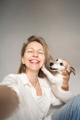 Happy friends selfie. blond woman and small dog Jack Russell terrier. woman smiling looking at camera and touching her face.  Dog owner and pet having fun in studio grey background. empty copy space