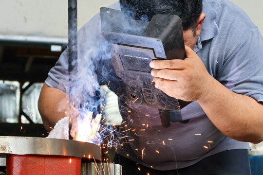 A professional welder working on a metal object, holding a protective helmet while arc welding produces bright light, blue smoke, and sparks in a workshop.