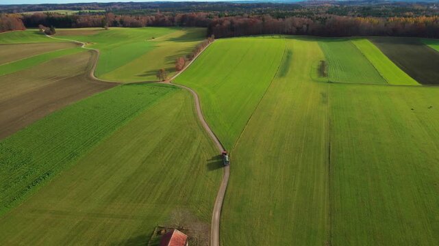 Aerial view of vibrant green farm fields, bisected by a meandering path, with a tractor visible near the road, Andechs, Bayern, Germany.
