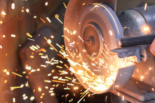 Close-up of a bench grinder in action, sharpening a metal rod and creating a shower of bright orange sparks and light in a dark mechanical workshop environment.