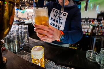 A close-up shot of a bartender's hands pouring a fresh, golden draft beer from a tap into a glass mug in a lively pub setting, capturing the foam and bubbles