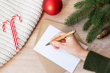 Female hand taking a pen. Mockup of an empty card,envelope on a wooden background, red ball, knitted blanket, christmas decor. New year and Christmas. Copy space.