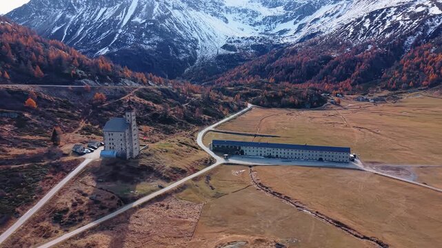 Aerial view of the Simplon Hospice nestled amidst brown fields and snow-capped mountains, contrasting with the clear sky, Simplon Hospice, Wallis, Switzerland.