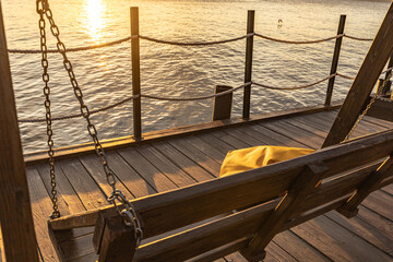 Golden sunset over the calm sea, a wooden pier and swing with a yellow rucksack. Travel, leisure, and seaside lifestyle.