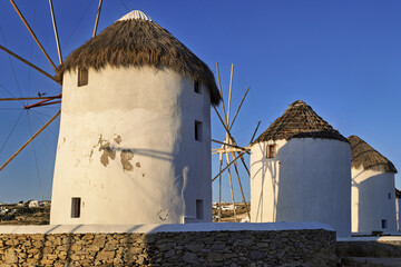 
Group of traditional windmills in Mykonos, Greece, with white cylindrical structures and conical thatched roofs, set on a stone platform under sunrise sky