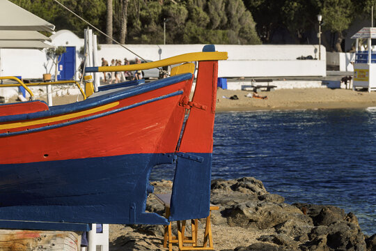 Brightly painted wooden boat in Mykonos resting on a rocky shoreline, featuring vivid red, blue, and yellow colors, with calm blue water and a sandy beach in the background, - Powered by Adobe