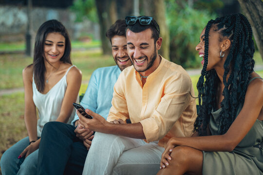 Diverse friends group laughing with a smartphone in the park. Happy multi-ethnic young adults sharing technology, concept of friendship and togetherness.
