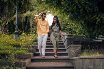 Happy diverse couple walking and smiling in a summer park. Multi-ethnic fashion lifestyle concept with two people on vacation on stone stairs.