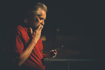 Older man in red shirt waiting for something and using mobile phone on night city location
