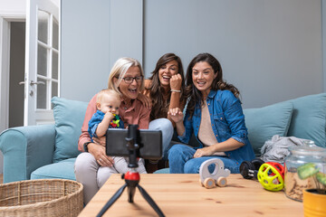 Three generations share a joyful video call—two women and their mother laugh while a baby plays on grandma’s lap; a smartphone tripod captures the scene as they connect with relatives online together