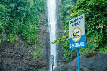 Indonesian. A warning sign with the Indonesian language "swimming is strictly prohibited" in red stands upright in front of a rushing waterfall as an advertising space.