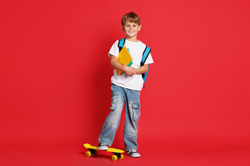 Cute boy with backpack, notebooks and penny board on red background