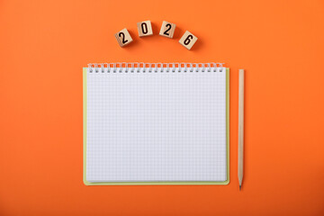 New 2026 year. Cubes with numbers, notebook and pencil on orange background, top view