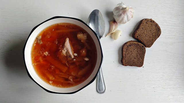 Red beetroot soup in bowl with rye Borodino bread, garlic. Russian simple rustic cuisine. Plate with first course on wooden table. Traditional Russian borscht, two bread pieces, garlic head and cloves