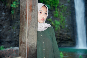 asian muslim woman with pensive expression standing behind wooden pole with waterfall view in...