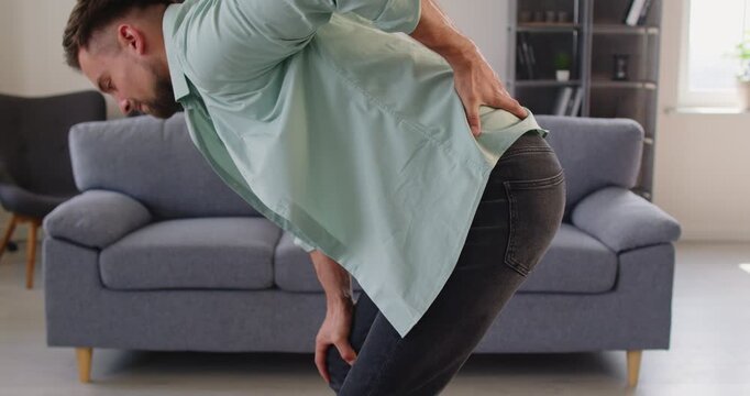 Young adult man at home bends forward, clutching lower back with pain and discomfort from backache, strain, injury, or sciatica affecting spine and posture. Man suffers acute lower back pain.