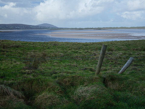 Windy Coastal Field in County Donegal Ireland With Wooden Fence Posts Overlooking Tidal Inlet Under Cloudy Sky