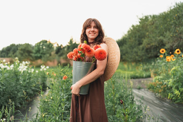 Woman smiling with a bucket of fresh dahlias on a rural farm during sunset
