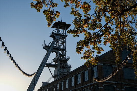Historic mining headframe silhouetted at sunset with industrial architecture, string lights, and autumn foliage in view.