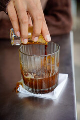 The moment a woman pours a strong espresso with golden crema into a glass filled with ice cubes. A close-up of the coffee being poured into a glass filled with ice cubes.
