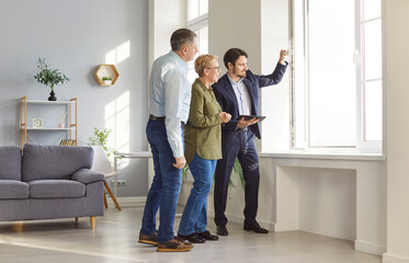 Male real estate agent showing senior couple stunning view from window in new house or apartment. Smiling elderly buyers evaluating property options and exploring opportunities for purchase or rent.