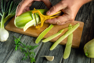 Hands skillfully peel fresh leeks on a wooden cutting board. Surrounding ingredients include green herbs and a clove of garlic, creating an inviting cooking atmosphere