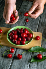 Hands are carefully holding cherries above a green bowl filled with freshly picked cherries on a wooden surface. Some cherries are scattered around, adding to the scene