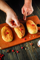 Hands are carefully placing red berries on freshly baked bread rolls on a wooden cutting board. Surrounding spices add warmth to the kitchen atmosphere