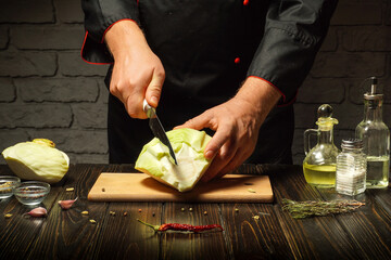 A skilled chef cuts a head of cabbage in a dimly lit kitchen. Ingredients are neatly arranged on the wooden cutting board, showcasing a culinary scene