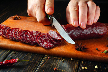 Hands carefully slice a piece of cured sausage into even rounds on a wooden cutting board, surrounded by colorful spices and herbs in a cozy kitchen setting