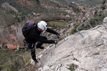 A climber ascends a steep rocky wall on a via ferrata in Castillo de Locub n, Ja n, Spain, using safety cables and metal steps surrounded by olive groves.