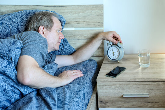 Lying in bed, a man reaches toward the alarm clock on his bedside table to switch it off, capturing a familiar moment of early morning routine and the challenge of waking up.
