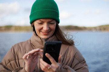 A smiling woman in a vibrant green beanie and tan puffer jacket uses her black smartphone outdoors by a calm blue lake under soft, diffused daylight.