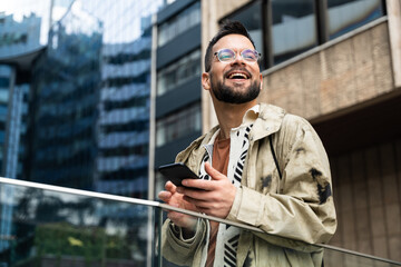 Young professional in a modern business district checks his phone—smiling at good news and managing messages, payments, maps, and schedules on the go while commuting between meetings across the city