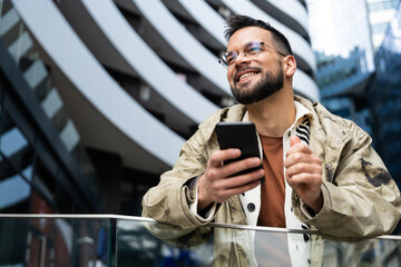 Young professional in a modern business district checks his phone—smiling at good news and managing messages, payments, maps, and schedules on the go while commuting between meetings across the city